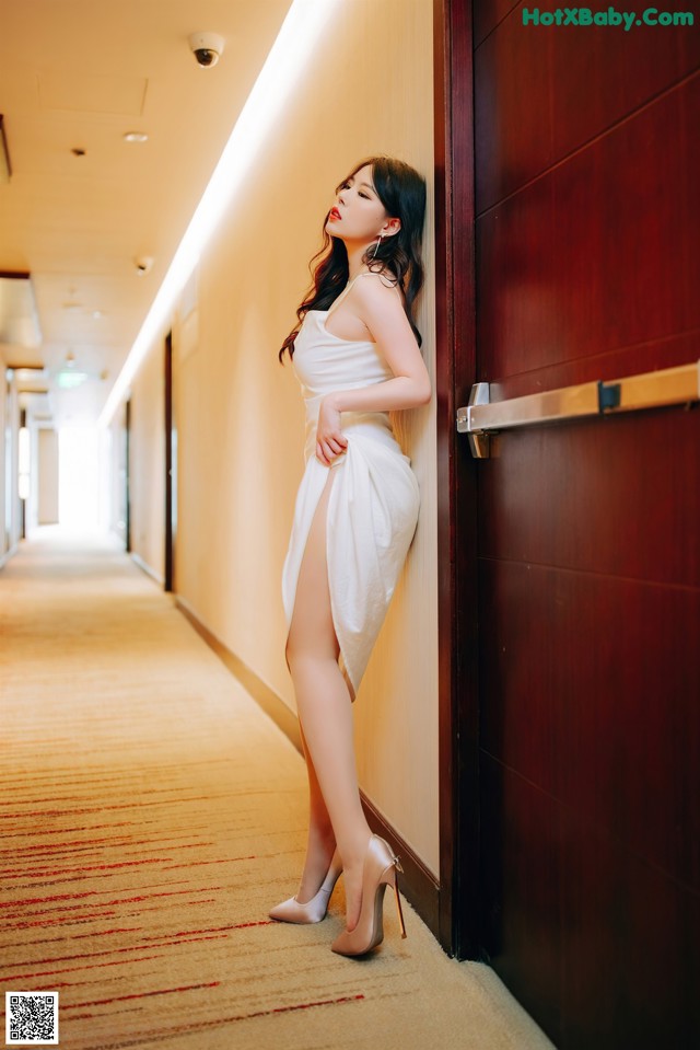 A woman in a white dress leaning against a wall.